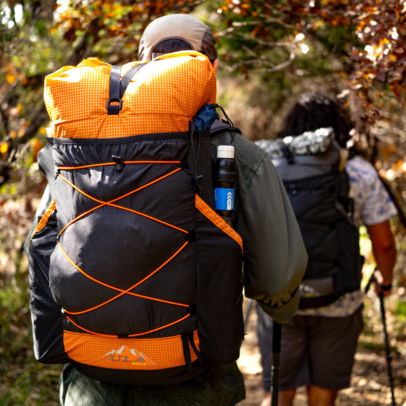 Two backpackers go hiking in the fall. The leaves are changing. In the foreground, one of the hikers wears a tangerine colored UltraGrid Nexus.