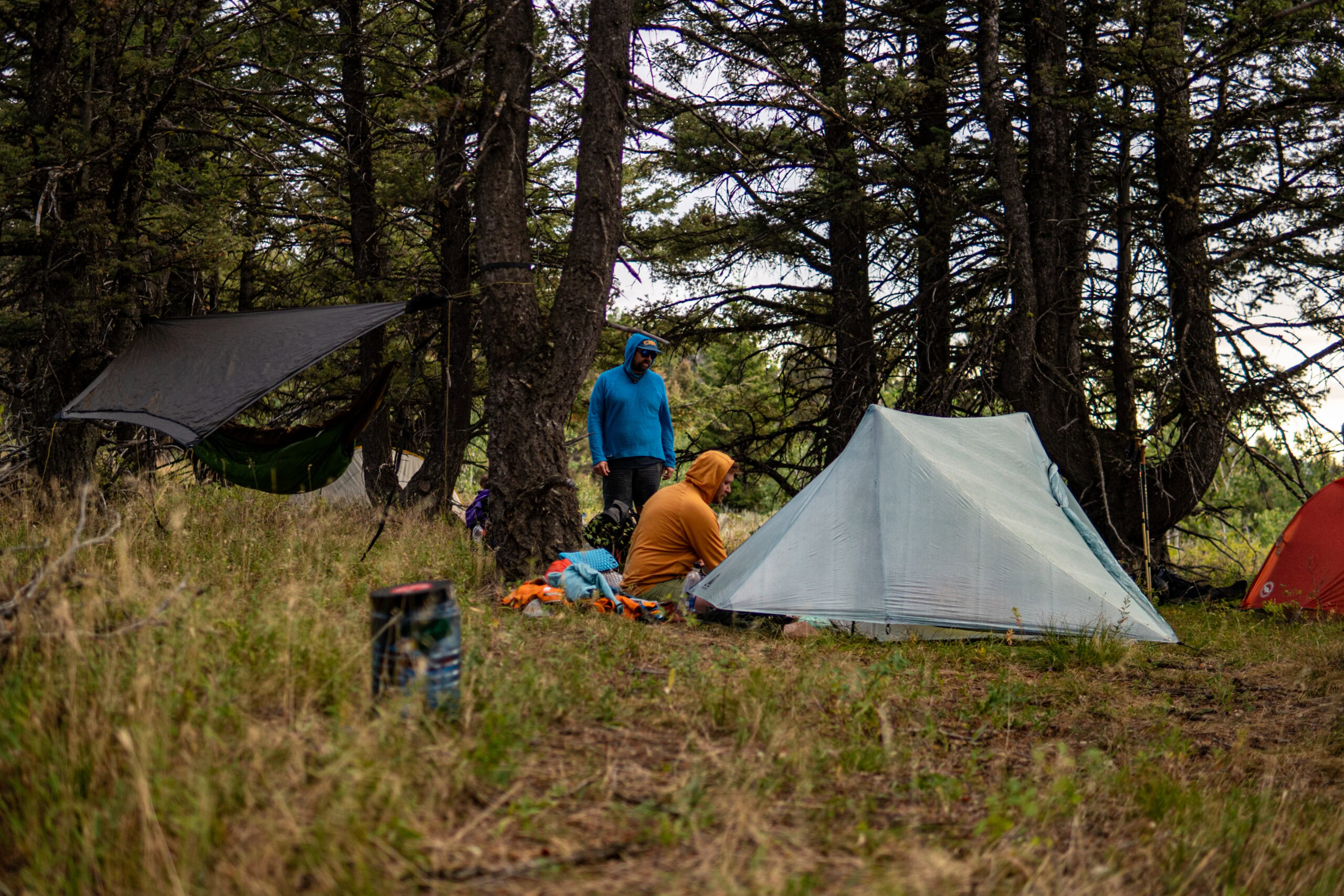 Two hikers sit outside their ultralight shelters. They are packing for departure. There is a bear canister in the foreground.